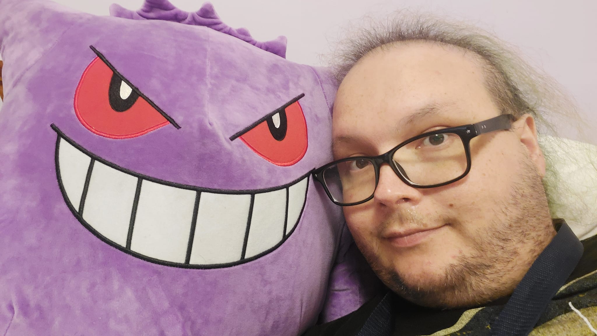 A man with thinning green hair lies in bed, holding a large Gengar plush